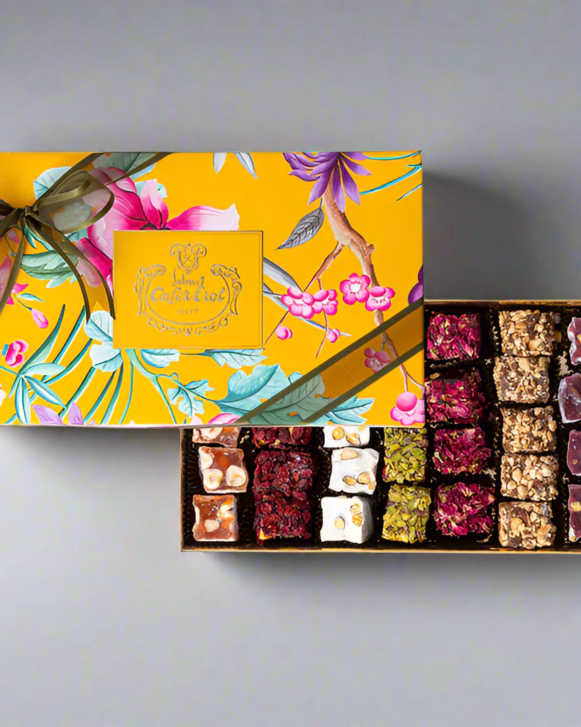 A decorative cardboard box of Mixed Roll Turkish Delights, with a floral pattern on the cover, and a variety of rolled sweets with different fillings visible through the transparent lid.